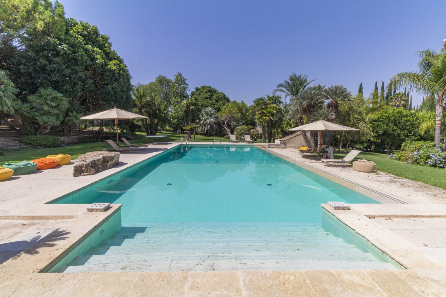 Beautiful outdoor swimming pool surrounded by lush greenery and palm trees, featuring comfortable lounge chairs and shaded umbrellas. The pool's crystal-clear water reflects the blue sky, creating a serene and inviting atmosphere for relaxation and leisure. Colorful bean bags add a fun touch to the outdoor space. | Only on SopranoVillas