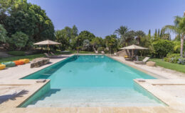 Beautiful outdoor swimming pool surrounded by lush greenery and palm trees, featuring comfortable lounge chairs and shaded umbrellas. The pool's crystal-clear water reflects the blue sky, creating a serene and inviting atmosphere for relaxation and leisure. Colorful bean bags add a fun touch to the outdoor space.
