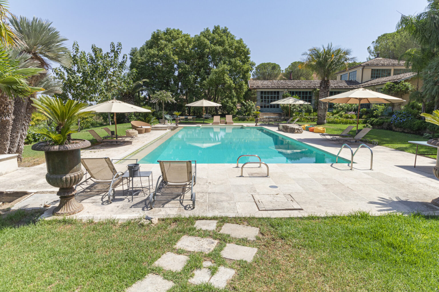 Luxurious backyard pool area featuring a sparkling blue pool, sun loungers, and shaded umbrellas surrounded by lush greenery and landscaped gardens. Perfect for relaxation and outdoor gatherings. | Only on SopranoVillas