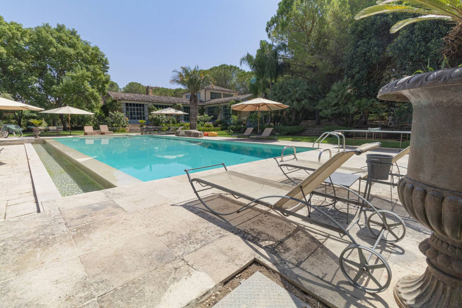 Luxurious outdoor pool area featuring a sparkling turquoise pool surrounded by sun loungers, umbrellas, and lush greenery, with a stylish home in the background. Ideal for relaxation and leisure. | Only on SopranoVillas