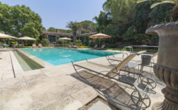 Luxurious outdoor pool area featuring a sparkling turquoise pool surrounded by sun loungers, umbrellas, and lush greenery, with a stylish home in the background. Ideal for relaxation and leisure.