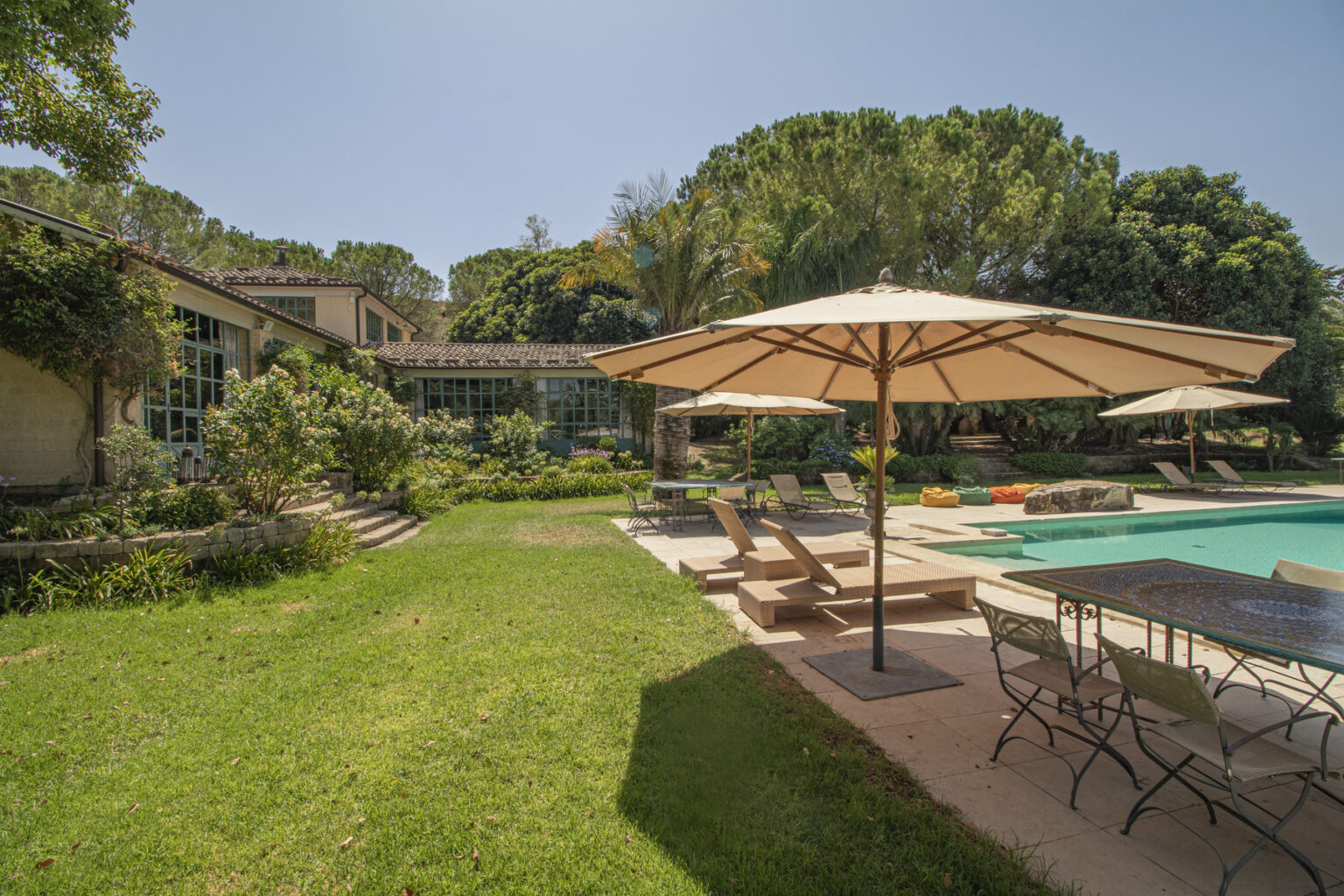 Luxurious backyard with a swimming pool, sun loungers, and a dining area under an umbrella, surrounded by lush greenery and a modern home. Ideal for outdoor relaxation and entertaining. | Only on SopranoVillas