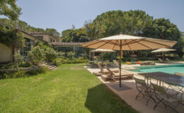 Luxurious backyard with a swimming pool, sun loungers, and a dining area under an umbrella, surrounded by lush greenery and a modern home. Ideal for outdoor relaxation and entertaining.