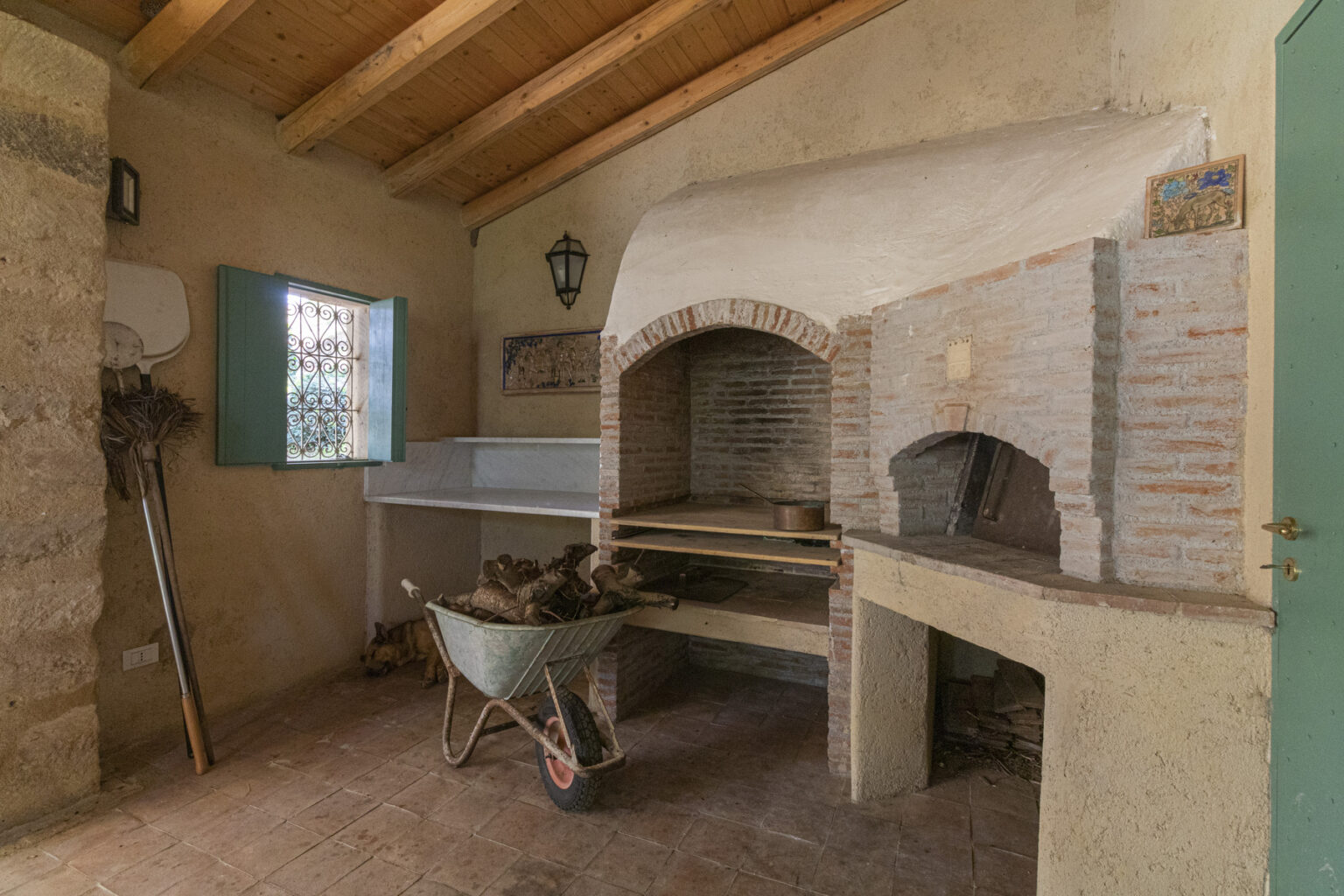 Rustic indoor pizza oven with a stone structure, wooden shelves, and a wheelbarrow for wood storage, set in a cozy kitchen environment with natural light and wooden beams. | Only on SopranoVillas