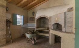 Rustic indoor pizza oven with a stone structure, wooden shelves, and a wheelbarrow for wood storage, set in a cozy kitchen environment with natural light and wooden beams.