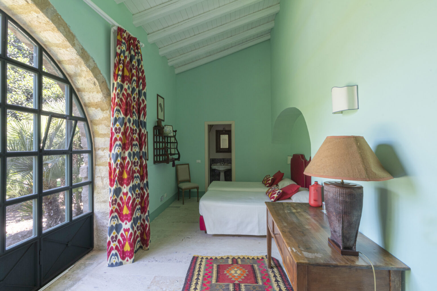 Bright and airy bedroom featuring a turquoise wall, large arched window, and colorful patterned curtains. The room includes a white bed with decorative pillows, a wooden nightstand with a lamp, and a traditional rug, creating a cozy and inviting atmosphere. | Only on SopranoVillas