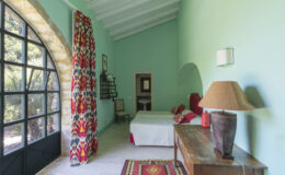 Bright and airy bedroom featuring a turquoise wall, large arched window, and colorful patterned curtains. The room includes a white bed with decorative pillows, a wooden nightstand with a lamp, and a traditional rug, creating a cozy and inviting atmosphere.