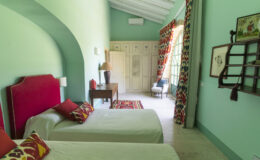 Bright and airy bedroom featuring two twin beds with colorful accent pillows, a wooden bedside table, and a decorative area rug. The room is painted in a soothing mint green, complemented by vibrant patterned curtains and natural light streaming through large windows. Stylish decor elements add a touch of charm to this inviting space.