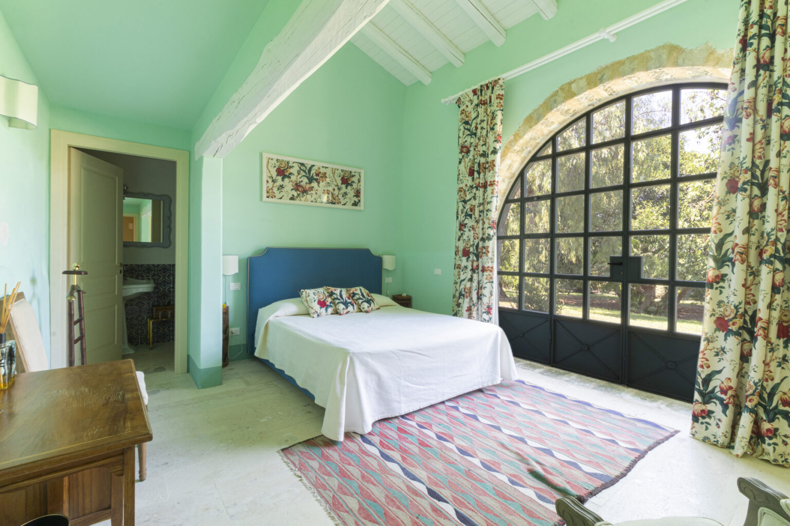 Bright and airy bedroom featuring a turquoise color scheme, a blue upholstered bed with decorative pillows, floral curtains, and a large arched window that offers natural light. A wooden dresser adds warmth to the space, complemented by a colorful area rug. | Only on SopranoVillas