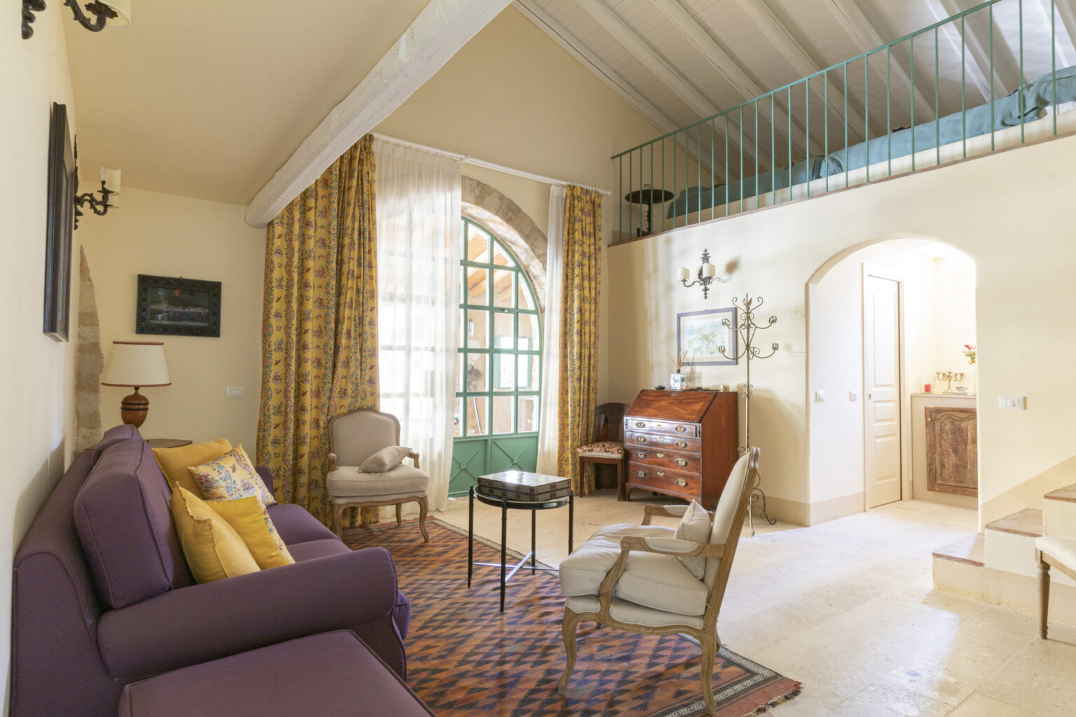 Cozy living room featuring a purple sofa with colorful cushions, elegant armchairs, and a wooden side table. Bright curtains frame a large window with a green door, while a vintage dresser and wall decor add charm. A lofted sleeping area is visible above, enhancing the spacious ambiance. | Only on SopranoVillas
