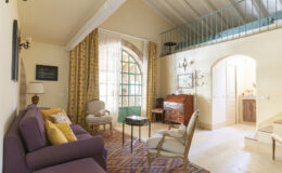 Cozy living room featuring a purple sofa with colorful cushions, elegant armchairs, and a wooden side table. Bright curtains frame a large window with a green door, while a vintage dresser and wall decor add charm. A lofted sleeping area is visible above, enhancing the spacious ambiance.