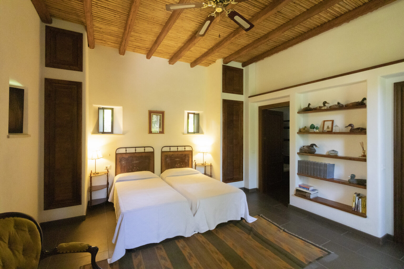 Cozy bedroom featuring two twin beds with white bedding, rustic wooden ceiling beams, warm lighting from bedside lamps, and a wooden shelf displaying decorative items. The room has a serene atmosphere and natural light from windows. | Only on SopranoVillas