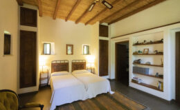 Cozy bedroom featuring two twin beds with white bedding, rustic wooden ceiling beams, warm lighting from bedside lamps, and a wooden shelf displaying decorative items. The room has a serene atmosphere and natural light from windows.
