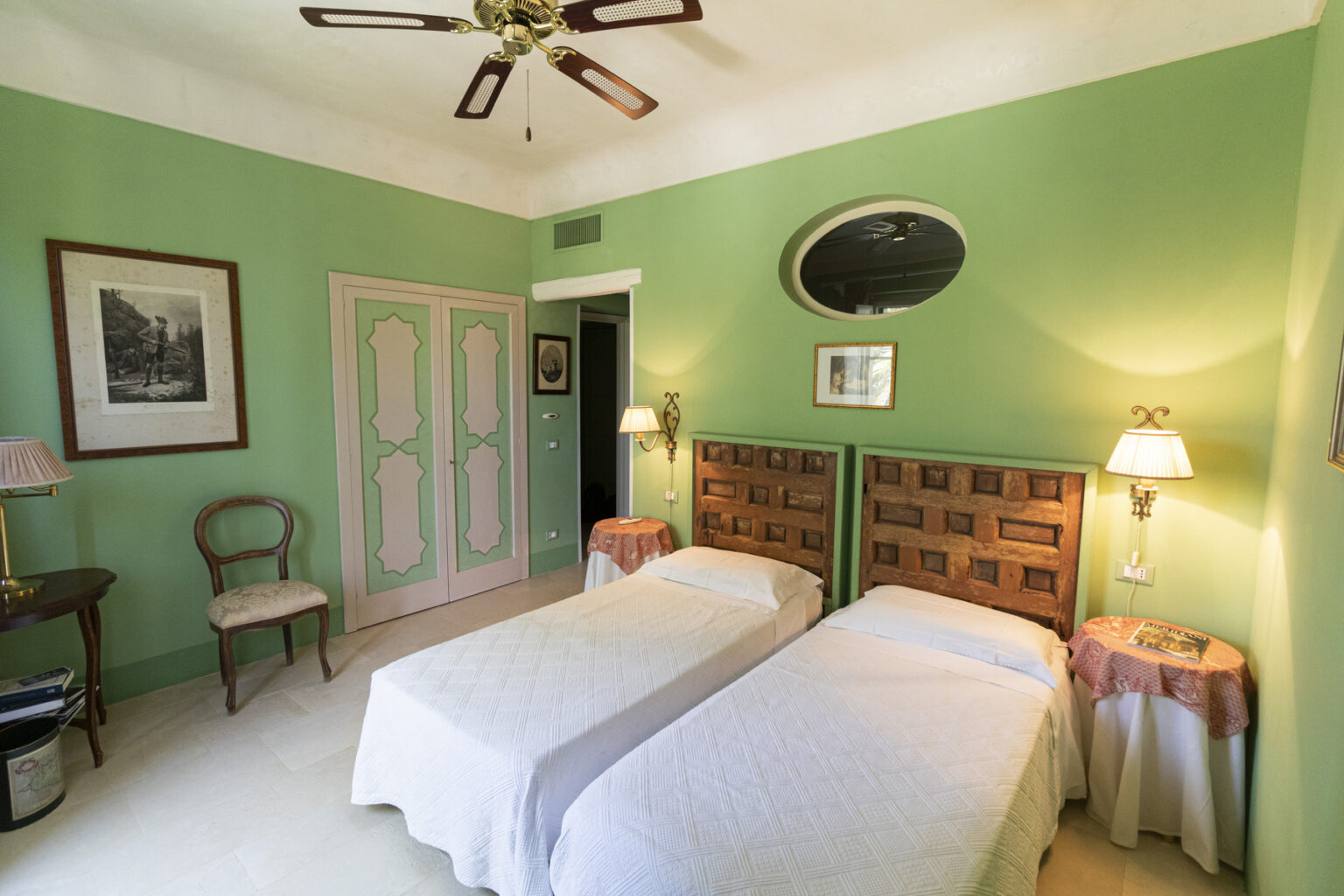 Bright and airy guest room featuring two twin beds with white bedding, green walls, vintage decor, and a ceiling fan. The room includes a small seating area with a chair, side tables, and framed artwork, creating a cozy and inviting atmosphere. | Only on SopranoVillas