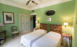 Bright and airy guest room featuring two twin beds with white bedding, green walls, vintage decor, and a ceiling fan. The room includes a small seating area with a chair, side tables, and framed artwork, creating a cozy and inviting atmosphere.