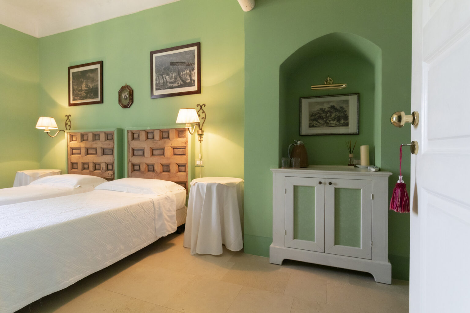 Cozy bedroom featuring two twin beds with patterned headboards, a green accent wall, vintage artwork, and a white cabinet with decorative items. The room is well-lit with bedside lamps, creating a warm and inviting atmosphere. | Only on SopranoVillas