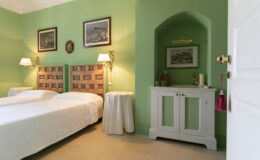 Cozy bedroom featuring two twin beds with patterned headboards, a green accent wall, vintage artwork, and a white cabinet with decorative items. The room is well-lit with bedside lamps, creating a warm and inviting atmosphere.