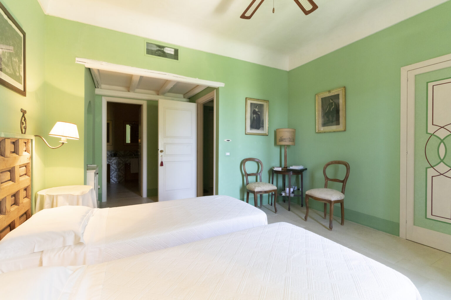 Bright and airy guest room featuring two twin beds, green walls, and vintage decor. The room includes a small seating area with two chairs, a bedside lamp, and artwork on the walls, creating a cozy and inviting atmosphere. | Only on SopranoVillas