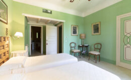 Bright and airy guest room featuring two twin beds, green walls, and vintage decor. The room includes a small seating area with two chairs, a bedside lamp, and artwork on the walls, creating a cozy and inviting atmosphere.