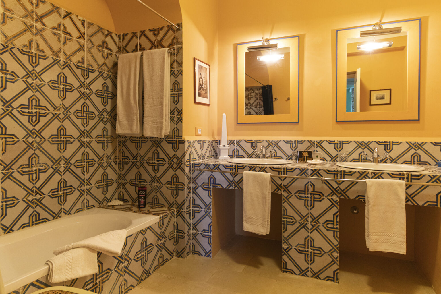 Brightly lit bathroom featuring a bathtub and dual sinks with stylish mirrors. The walls are adorned with colorful patterned tiles, complemented by white towels hanging on a rack. The warm orange hue adds a cozy atmosphere to the space. | Only on SopranoVillas