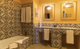 Brightly lit bathroom featuring a bathtub and dual sinks with stylish mirrors. The walls are adorned with colorful patterned tiles, complemented by white towels hanging on a rack. The warm orange hue adds a cozy atmosphere to the space.