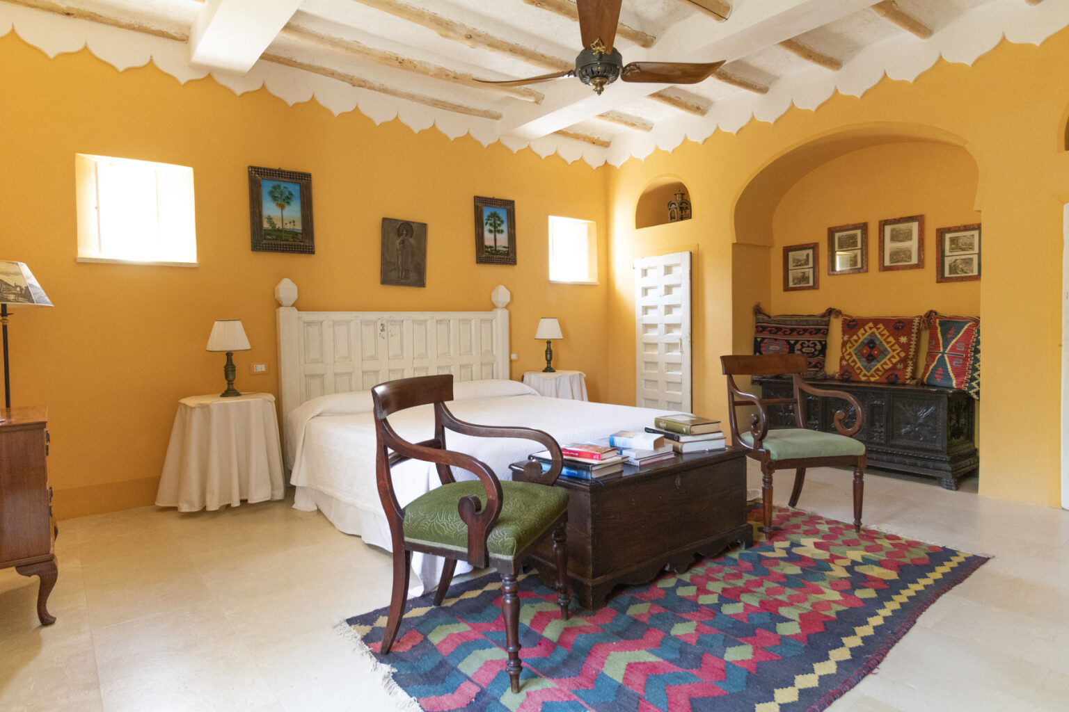 Cozy bedroom featuring a king-sized bed, antique wooden furniture, and vibrant orange walls. The room includes a colorful area rug, a ceiling fan, and framed artwork, creating a warm and inviting atmosphere. | Only on SopranoVillas