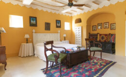 Cozy bedroom featuring a king-sized bed, antique wooden furniture, and vibrant orange walls. The room includes a colorful area rug, a ceiling fan, and framed artwork, creating a warm and inviting atmosphere.