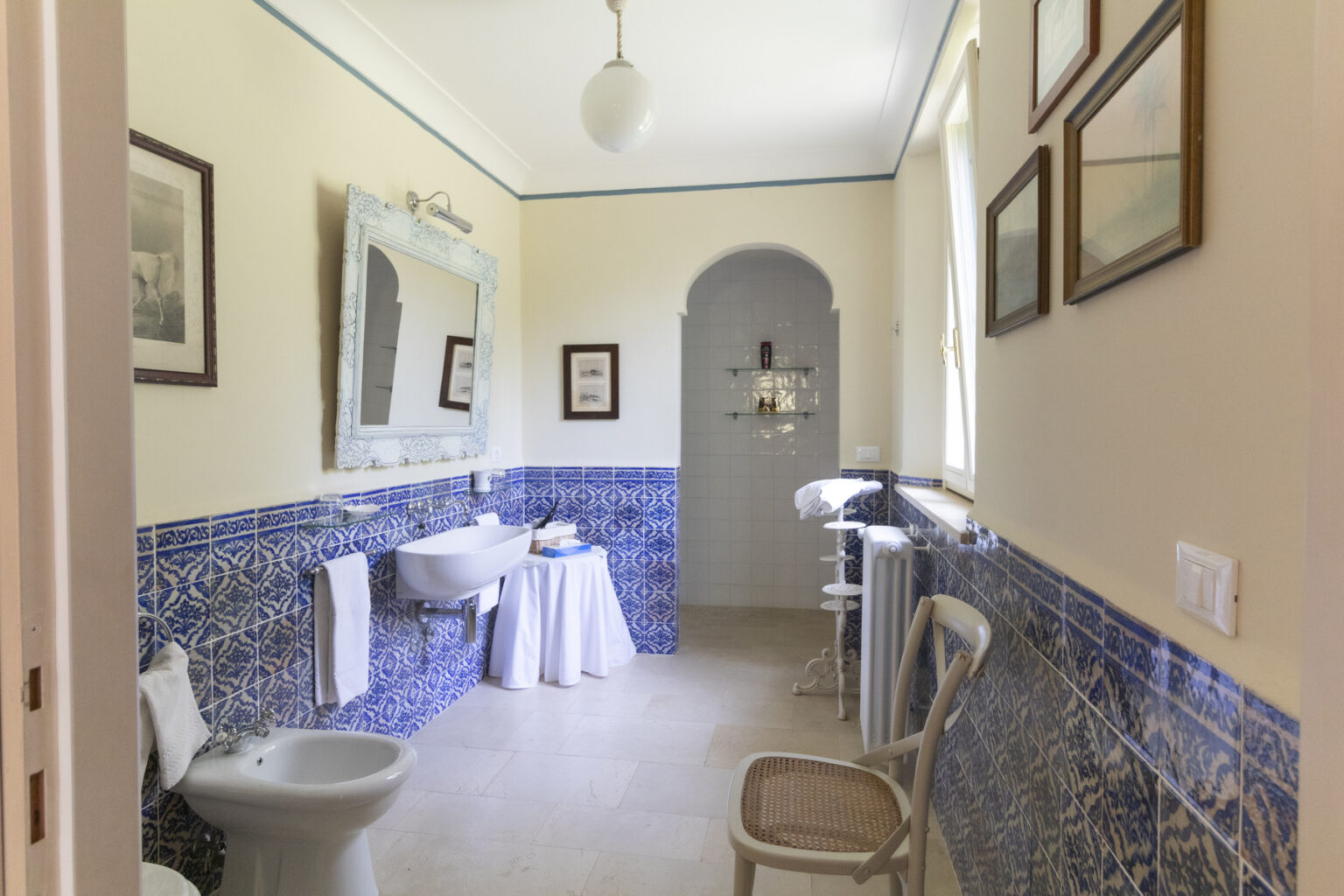 Bright and airy bathroom featuring blue tiled walls, a vintage mirror, and framed artwork. The space includes a white sink, a toilet, and a small table with towels, creating a charming and inviting atmosphere. | Only on SopranoVillas