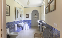 Bright and airy bathroom featuring blue tiled walls, a vintage mirror, and framed artwork. The space includes a white sink, a toilet, and a small table with towels, creating a charming and inviting atmosphere.