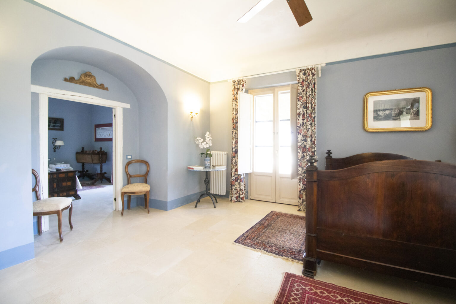 Interior view of a cozy bedroom featuring a wooden bed, light blue walls, patterned curtains, and a vintage chair. The room has a warm ambiance with natural light streaming through the window, showcasing a classic design aesthetic. | Only on SopranoVillas