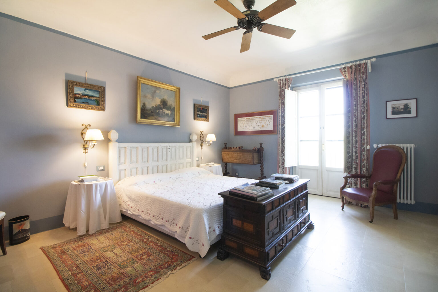 Cozy bedroom featuring a white bed with decorative pillows, wooden furniture, and soft blue walls. Natural light streams through large windows, highlighting a vintage rug and artwork on the walls, creating a warm and inviting atmosphere. | Only on SopranoVillas