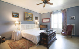 Cozy bedroom featuring a white bed with decorative pillows, wooden furniture, and soft blue walls. Natural light streams through large windows, highlighting a vintage rug and artwork on the walls, creating a warm and inviting atmosphere.