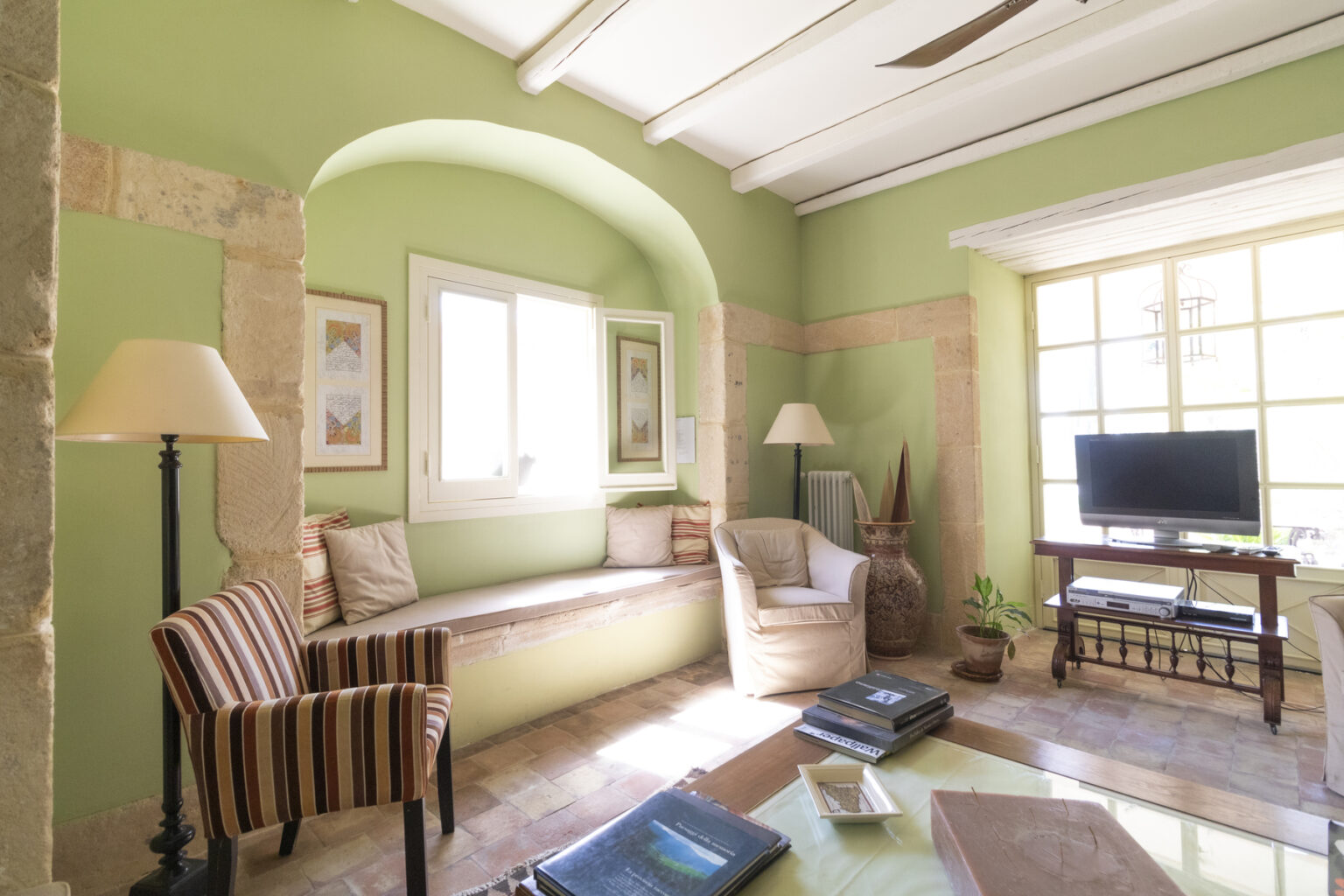 Cozy living room featuring green walls, natural light from large windows, a comfortable seating area with striped chairs, and a TV on a wooden stand, complemented by decorative plants and artwork. | Only on SopranoVillas