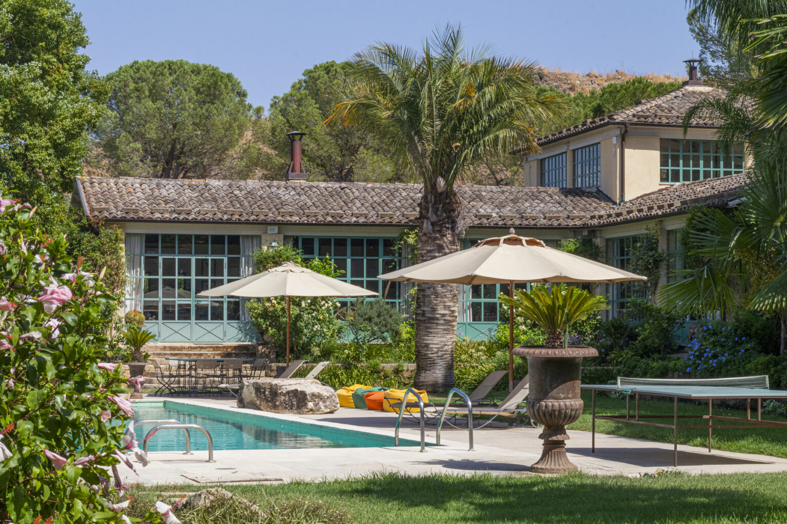 Luxurious villa with a swimming pool surrounded by lush greenery, featuring sun umbrellas and a stone fountain, ideal for relaxation and outdoor living. | Only on SopranoVillas