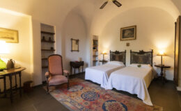 Cozy bedroom featuring two twin beds, a vintage armchair, and warm lighting, with an elegant rug and arched ceiling design. Ideal for relaxation and comfort.