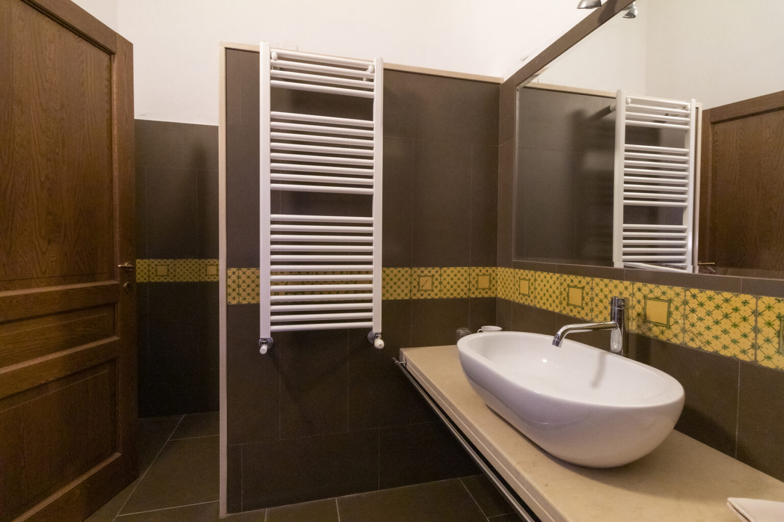 Modern bathroom interior featuring a sleek white bathtub, stylish wall-mounted towel warmer, and elegant mirror. The design incorporates dark wood accents and a decorative yellow tile border, creating a warm and inviting atmosphere. | Only on SopranoVillas