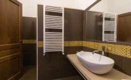 Modern bathroom interior featuring a sleek white bathtub, stylish wall-mounted towel warmer, and elegant mirror. The design incorporates dark wood accents and a decorative yellow tile border, creating a warm and inviting atmosphere.