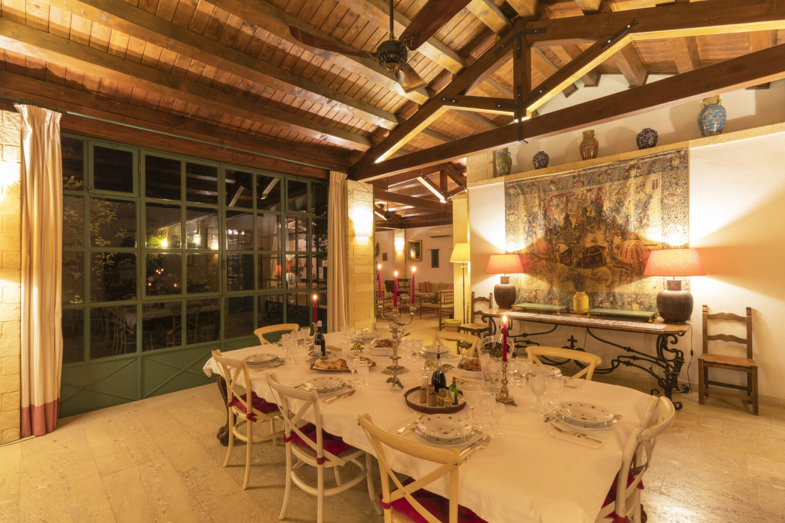 Cozy dining room with a large table set for a meal, featuring elegant tableware, candles, and a vintage tapestry on the wall. The space has wooden beams, soft lighting, and a large window that opens to a garden, creating an inviting atmosphere for gatherings. | Only on SopranoVillas