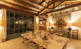 Cozy dining room with a large table set for a meal, featuring elegant tableware, candles, and a vintage tapestry on the wall. The space has wooden beams, soft lighting, and a large window that opens to a garden, creating an inviting atmosphere for gatherings.