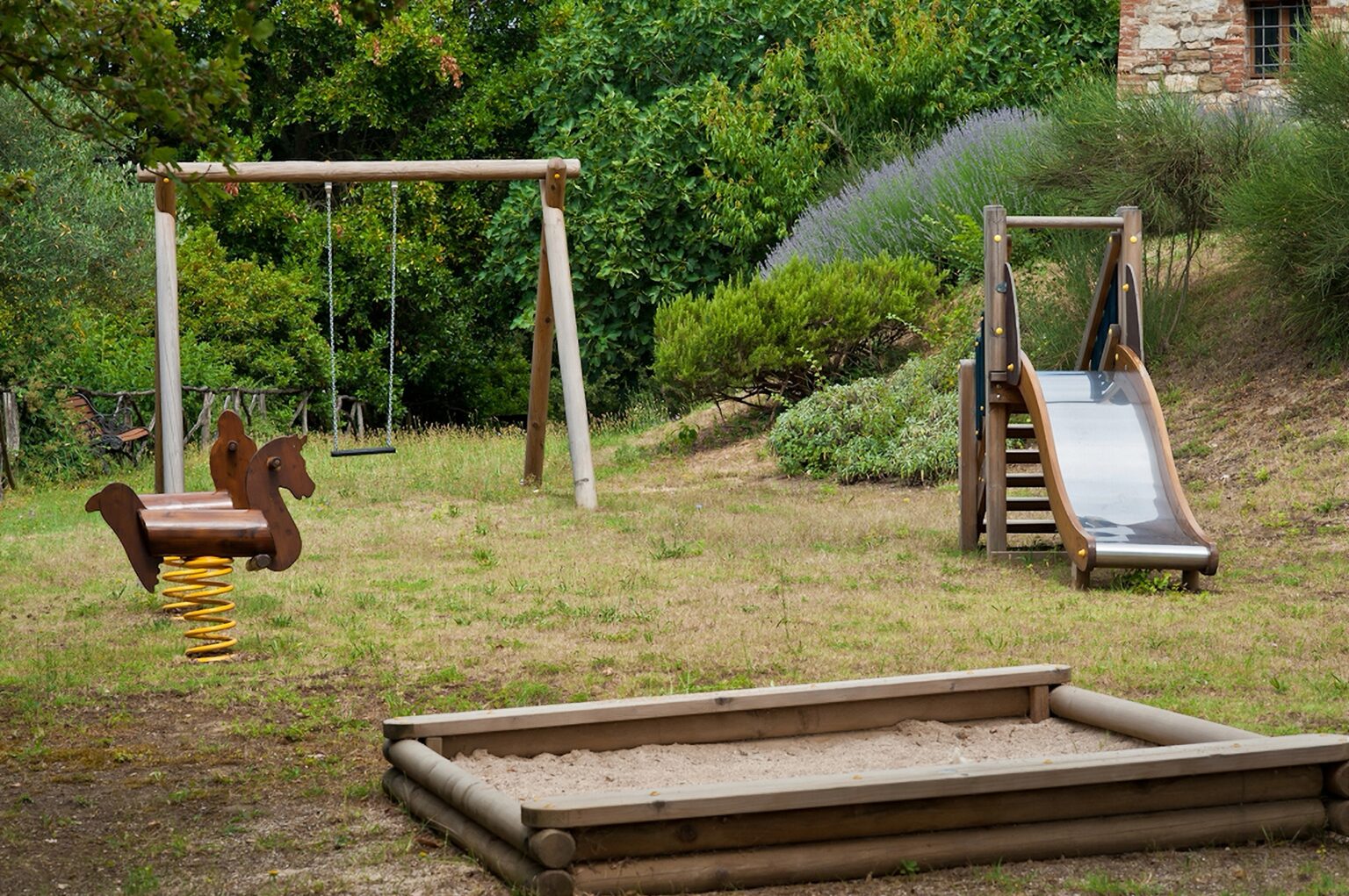 Playground equipment in a natural setting featuring a wooden swing set, a spring rider designed as a horse, a slide, and a sand pit, surrounded by greenery and shrubs. Ideal for children's outdoor play and recreation. | Only on SopranoVillas