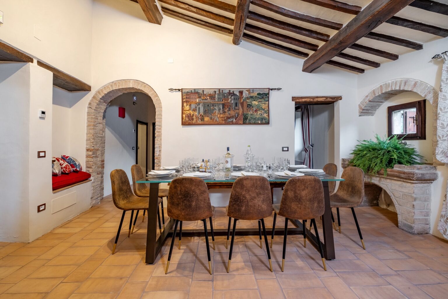 Elegant dining room featuring a large glass-topped table surrounded by stylish brown chairs, set for a meal with fine dishware. The room boasts rustic wooden beams and a charming mural on the wall, complemented by a cozy nook with colorful cushions and greenery. | Only on SopranoVillas