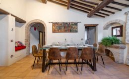 Elegant dining room featuring a large glass-topped table surrounded by stylish brown chairs, set for a meal with fine dishware. The room boasts rustic wooden beams and a charming mural on the wall, complemented by a cozy nook with colorful cushions and greenery.