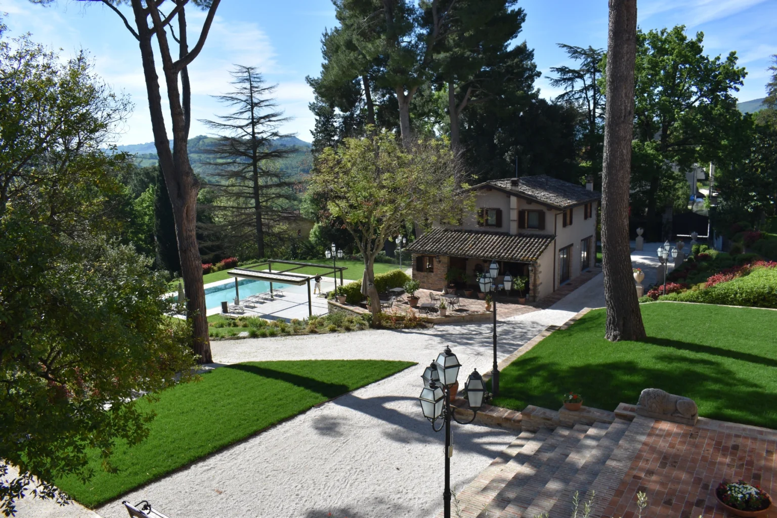 Scenic view of a landscaped garden featuring a swimming pool, a charming house with a rustic roof, and lush greenery. The image captures outdoor seating areas and decorative lighting, set against a backdrop of trees and rolling hills. Ideal for showcasing outdoor living spaces and tranquil retreats. | Only on SopranoVillas
