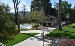 Scenic view of a landscaped garden featuring a swimming pool, a charming house with a rustic roof, and lush greenery. The image captures outdoor seating areas and decorative lighting, set against a backdrop of trees and rolling hills. Ideal for showcasing outdoor living spaces and tranquil retreats.
