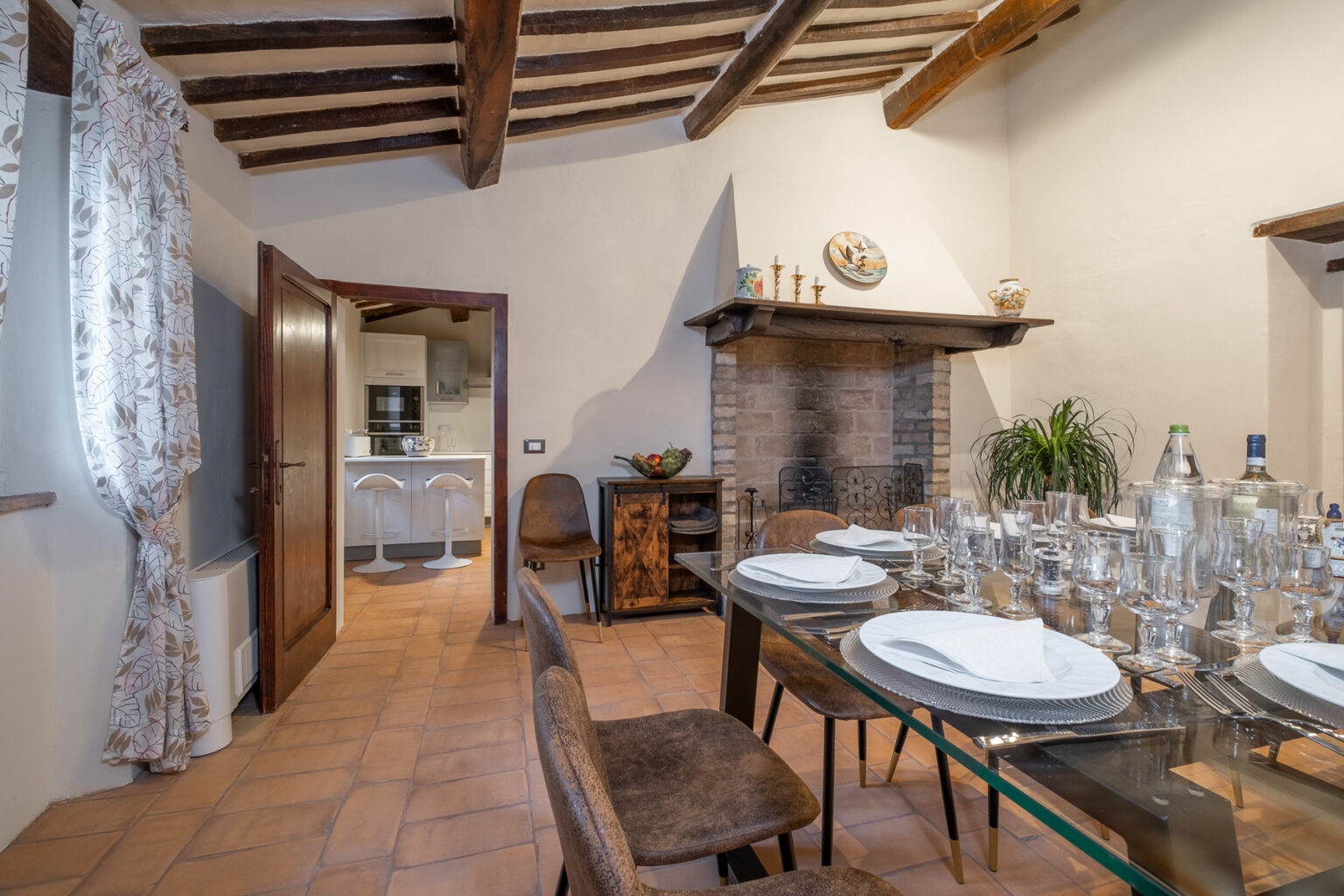 Cozy dining area featuring a glass table set for a meal, surrounded by stylish chairs, with a rustic fireplace and a modern kitchen visible in the background. The room showcases wooden beams and warm tile flooring, creating an inviting atmosphere. | Only on SopranoVillas