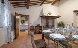 Cozy dining area featuring a glass table set for a meal, surrounded by stylish chairs, with a rustic fireplace and a modern kitchen visible in the background. The room showcases wooden beams and warm tile flooring, creating an inviting atmosphere.