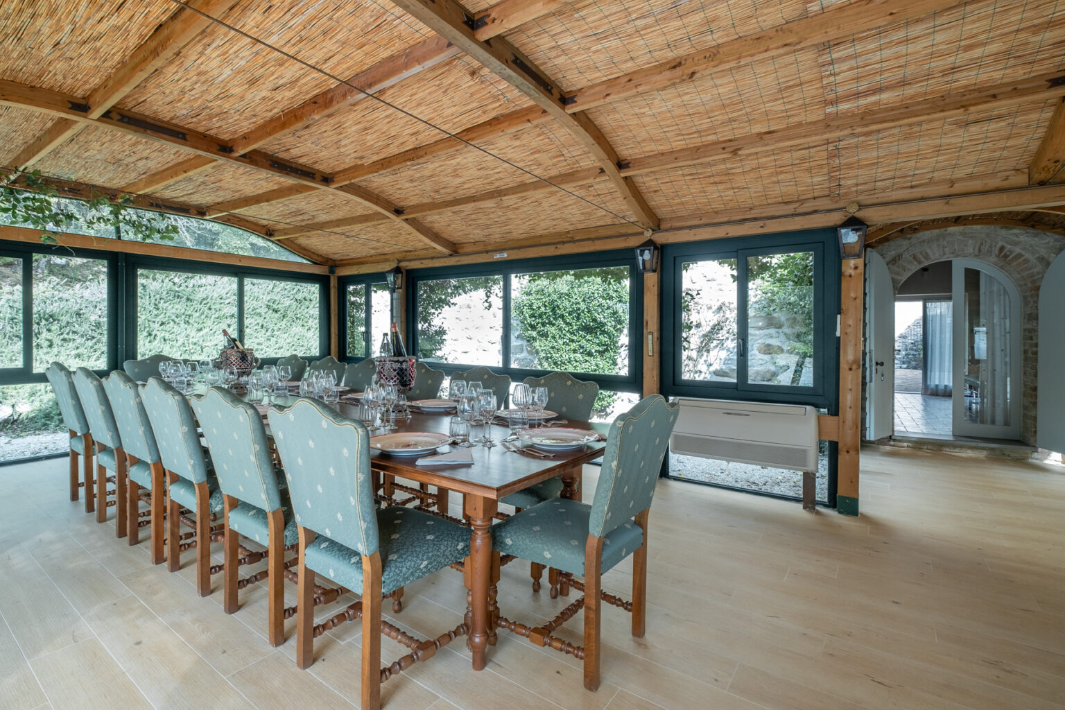 Spacious dining area with a large wooden table set for a meal, featuring elegant glassware and decorative plates. Surrounded by large windows, the room has a warm wooden ceiling and a cozy atmosphere, ideal for gatherings. | Only on SopranoVillas