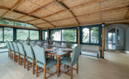 Spacious dining area with a large wooden table set for a meal, featuring elegant glassware and decorative plates. Surrounded by large windows, the room has a warm wooden ceiling and a cozy atmosphere, ideal for gatherings.