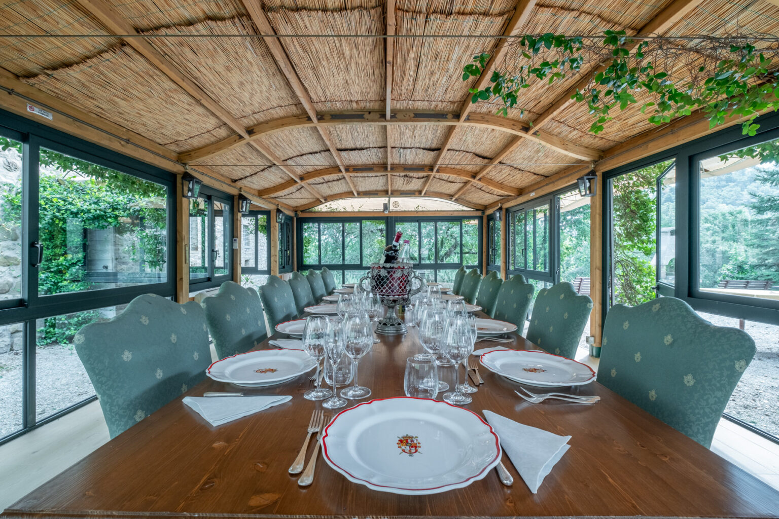 Elegant dining room with a long wooden table set for a formal meal, featuring fine china and crystal glassware. The room is adorned with a thatched roof and large windows, offering views of greenery outside, creating a serene atmosphere for gatherings. | Only on SopranoVillas
