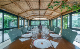 Elegant dining room with a long wooden table set for a formal meal, featuring fine china and crystal glassware. The room is adorned with a thatched roof and large windows, offering views of greenery outside, creating a serene atmosphere for gatherings.